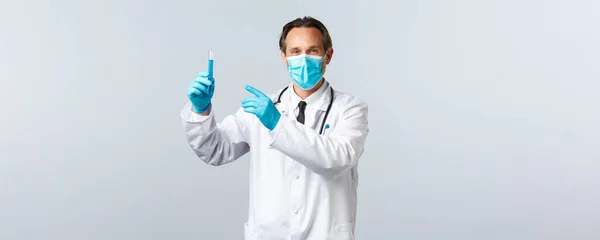 Covid-19: 바이러스 예방, 의료 종사자 및 예방 개념. 의료 마스크를 쓴 만족 스러운 미소 의사와 백신 샘플이 들어 있는 시험관을 가리키는 장갑을 낀 의사, 약품 승인 — 스톡 사진