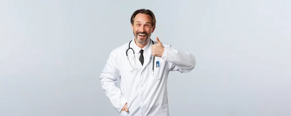 Covid-19, Coronavirus-Ausbruch, Gesundheitspersonal und Pandemiekonzept. Glücklich lächelnder Arzt im weißen Mantel lächelt zufrieden, zeigt Daumen hoch, empfiehlt und fördert Klinikdienstleistungen — Stockfoto