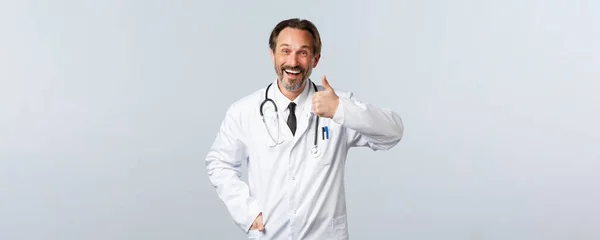 Covid-19, coronavirus outbreak, healthcare workers and pandemic concept. Happy smiling male doctor in white coat smiling pleased, show thumbs-up, recommend and promote clinic services — Stock Photo, Image