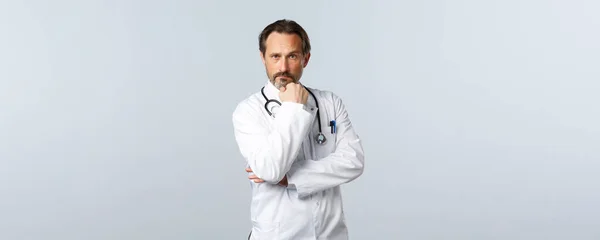 Covid-19, Coronavirus-Ausbruch, Gesundheitspersonal und Pandemiekonzept. Seriös aussehender männlicher Arzt in weißem Mantel, Berührungsbart und nachdenklicher Kamera, der Patienten behandelt — Stockfoto