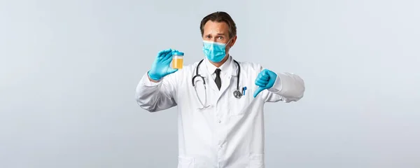 Covid-19: 바이러스 예방, 의료 종사자 및 예방 개념. 의료용 마스크와 장갑을 끼고 있는 정신과 의사는 소변 샘플을 보여 주고, 엄지손가락을 내리는 것은 안 좋은 검사 결과를 보여 주고, 하얀 배경을 보여 줍니다. — 스톡 사진