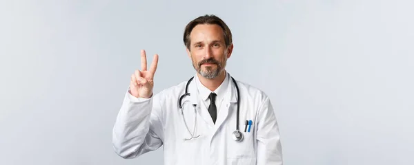 Covid-19, Prävention von Viren, Gesundheitspersonal und Impfkonzept. Optimistisch lächelnder Arzt in weißem Mantel zeigt V-Zeichen Frieden, ist begeistert, behandelt kranke Patienten, glaubt an gutes Ergebnis — Stockfoto