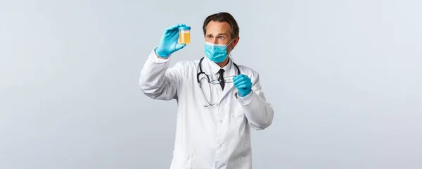 Covid-19, prevención de virus, trabajadores sanitarios y concepto de vacunación. Médico serio en máscara médica y guantes examinar la muestra de orina, mirar reflexivo y concentrado —  Fotos de Stock