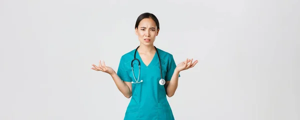 Ковід-19, медичні працівники, пандемія. Збентежена і розчарована азіатська медсестра, жінка-лікар в скрабах розтягнула руки вбік і відгинаючись турбує, може зрозуміти, чому — стокове фото