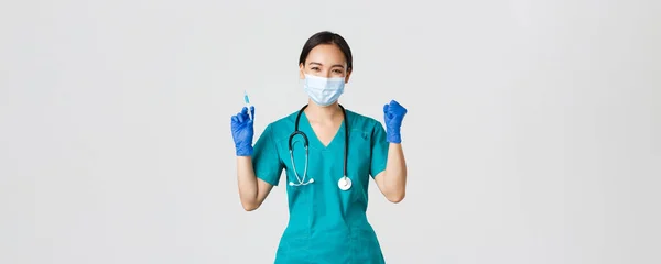 Covid-19, Coronavirus-Krankheit, Konzept des Gesundheitspersonals. Fröhlich triumphierende asiatische Ärztin, Ärztin in medizinischer Maske und Handschuhen, jubelt, während sie eine Spritze mit Impfstoff in der Hand hält — Stockfoto