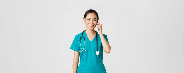 Covid-19, vårdpersonal och förebyggande av virus. Leende glad asiatisk kvinnlig läkare, ung praktikant i skurar talar i telefon, tittar på kameran hoppfull, vit bakgrund — Stockfoto