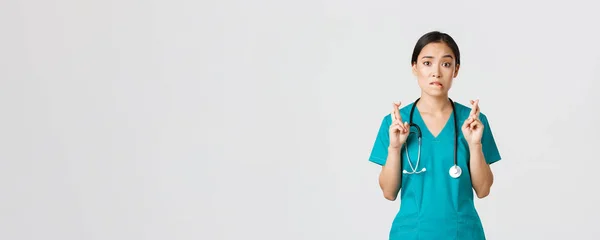 Covid-19, personnel de santé, concept pandémique. Espérant nerveuse asiatique femme stagiaire dans les gommages en attente de nouvelles, médecin croix doigts et mordre lèvre comme regarder la caméra, prier — Photo