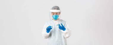 Covid-19, koronavirüs hastalığı, sağlık çalışanları konsepti. Ciddi görünümlü kadın hekim, kişisel koruma ekipmanlarında Asyalı doktor aşıyla birlikte ampul içine şırınga yerleştirin.