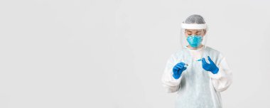 Covid-19, koronavirüs hastalığı, sağlık çalışanları konsepti. Ciddi görünümlü kadın hekim, kişisel koruma ekipmanlarında Asyalı doktor aşıyla birlikte ampul içine şırınga yerleştirin.
