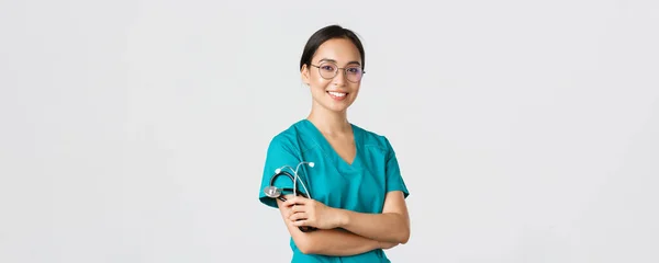 Covid-19, boala coronavirus, conceptul lucrătorilor din domeniul sănătății. Profesional medic asiatic arătos, lucrător medical în ochelari și scrubs, brațe încrucișate și zâmbitor, fundal alb — Fotografie, imagine de stoc