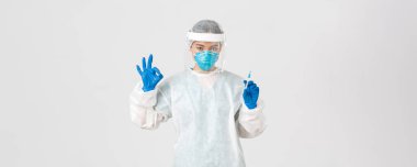 Covid-19, koronavirüs hastalığı, sağlık çalışanları konsepti. Kişisel koruma ekipmanlarında kendine güvenen ciddi Asyalı kadın doktor, iyi bir jest göster, aşıyla şırıngayı tut, beyaz arka plan