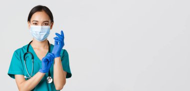 Covid-19, koronavirüs hastalığı, sağlık çalışanları konsepti. Profesyonel gülümseyen Asyalı kadın doktor, tıbbi maske ve önlüklü doktor, lastik eldiven giyin ro muayene, beyaz arka plan