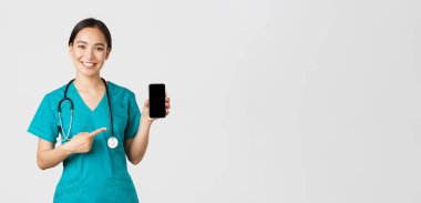 Covid-19, sağlık çalışanları ve online tıp konsepti. Gülümseyen Asyalı kadın doktor, ameliyat önlüklü doktor, hareket halindeki ekranı işaret ediyor, check-up 'ı indirmeyi, danışmanlık uygulamasını öneriyor