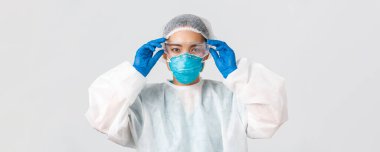 Covid-19, koronavirüs hastalığı, sağlık çalışanları konsepti. Ciddi görünümlü Asyalı kadın doktor, kişisel koruyucu ekipman, laboratuvara girmeden önce cam takmış, beyaz arka plan.
