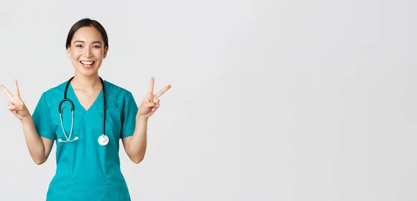 Covid-19, profissionais de saúde, conceito pandémico. feliz ásia feminino médico, enfermeira no scrubs mostrando paz kawaii gesto e sorrindo, permanecendo positivo durante o trabalho no hospital, branco fundo — Fotografia de Stock