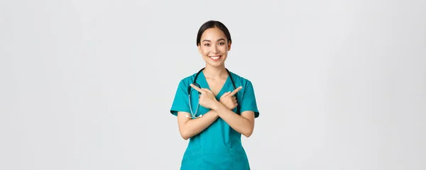 Covid-19, egészségügyi dolgozók, világjárvány. Mosolygó gyönyörű ázsiai nővér, gyakornok vagy orvos köpenyben mutatva oldalra, és a kamera, mutatja változatok, két választás, fehér háttér — Stock Fotó