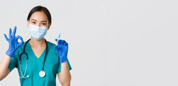 Covid-19, onemocnění koronavirem, koncept zdravotnických pracovníků. Roztomilý asijský lékař, lékař v lékařské masce a gumové rukavice ukazují v pořádku gesto a držet stříkačku s vakcínou, bílé pozadí — Stock fotografie