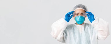 Covid-19, koronavirüs hastalığı, sağlık çalışanları konsepti. Ciddi görünümlü Asyalı kadın doktor, kişisel koruyucu ekipman, laboratuvara girmeden önce cam takmış, beyaz arka plan.