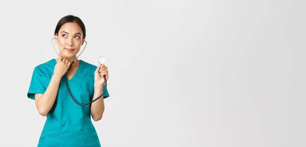 Covid-19, healthcare workers and preventing virus concept. Cute smiling asian female intern learn how listen lungs with stethscope, doctor examine patient, looking thoughtful upper left corner — Stock Photo, Image