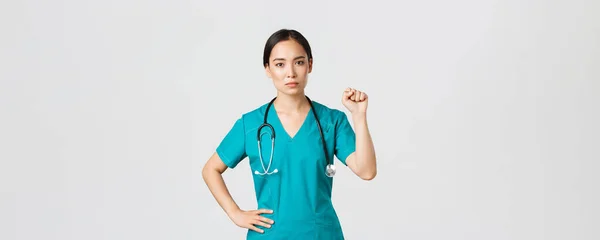Covid-19, egészségügyi dolgozók, világjárvány. Súlyos kinézetű, magabiztos, komoly ázsiai női orvos, aki támogatja kollégáit a koronavírus alatt, felemelve az öklét egységesen gesztusként — Stock Fotó
