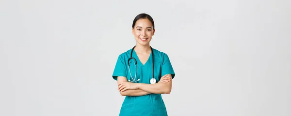Covid-19, pracownicy służby zdrowia, koncepcja pandemii. Portret pewnej siebie uśmiechniętej, atrakcyjnej azjatyckiej pielęgniarki w fartuchu, ze stetoskopem, klatką piersiową krzyżową i patrząc w kamerę, białe tło — Zdjęcie stockowe