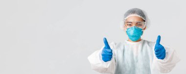 Covid-19, koronavirüs hastalığı, sağlık çalışanları konsepti. Şahsi koruma ekipmanlarıyla kendine güvenen Asyalı kadın doktorların yakın plan fotoğrafları. Başparmak kaldırma, tavsiye etme veya kalite güvencesi