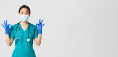 Covid-19, koronavirüs hastalığı, sağlık çalışanları konsepti. Etkilenmiş Asyalı kadın doktor, tıbbi maskeli doktor, kauçuk küreler ve önlükler. Tamam, jest, iltifat, övgü, mükemmel iş.