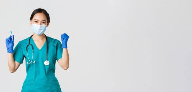 Covid-19, koronavirüs hastalığı, sağlık çalışanları konsepti. Neşeli zafer kazanan Asyalı kadın doktor, tıbbi maske ve eldiven takan doktor, aşıyla şırınga tutarken seviniyor.