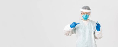 Covid-19, koronavirüs hastalığı, sağlık çalışanları konsepti. Ciddi bir Asyalı kadın doktor, PPE kişisel koruma ekipmanı doktoru, aşı şırıngasını işaret eden, beyaz arka plan