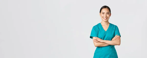 Ковид-19, медицинские работники, пандемия. Уверенный улыбающийся азиатский врач, женщина-медсестра в униформе стоя решительно, скрещенные руки груди на белом фоне. Врач готов к смене в клинике — стоковое фото