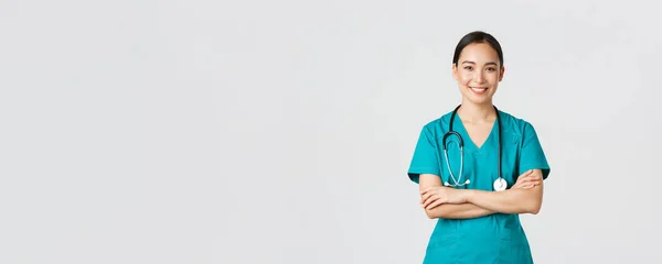 Covid-19, profissionais de saúde, conceito pandémico. Retrato de confiante sorrindo, atraente asiático enfermeira feminina em esfrega, com estetoscópio, cruz braços peito e olhando para a câmera, fundo branco — Fotografia de Stock