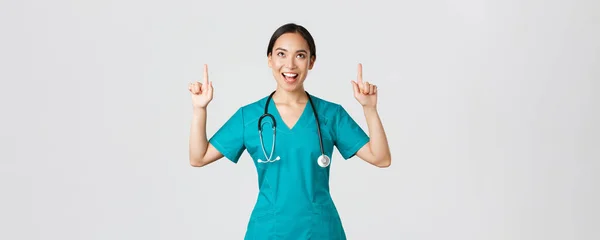 Covid-19, trabajadores de la salud, concepto pandémico. divertido alegre asiático mujer enfermera, médico o cirujano en scrubs apuntando dedos y mirando arriba con satisfecha cara, de pie blanco fondo — Foto de Stock