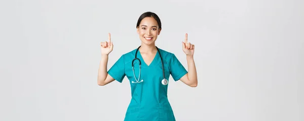 Ковид-19, медицинские работники, пандемия. Счастливая улыбающаяся азиатка-врач, медсестра в халате, показывающая рекламу, делающая объявление. Врач указывает пальцем на баннер — стоковое фото
