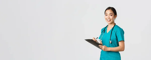 stock image Healthcare workers, preventing virus, quarantine campaign concept. Smiling cute asian female nurse, intern in scrubs, writing on clipboard, fill-in papers, looking a camera pleased, white background