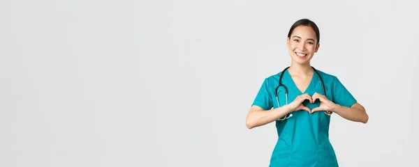 Ковид-19, медицинские работники, пандемия. Прекрасный заботливый азиатский врач, женщина-медсестра в скрабах показывает сердечный жест и улыбается, заботясь о пациентах с любовью, белый фон — стоковое фото