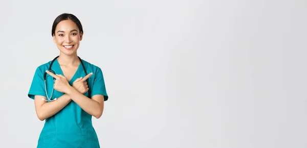 Covid-19, sjukvårdspersonal, pandemikoncept. Leende vacker asiatisk sjuksköterska, praktikant eller läkare i skurar pekar åt sidan och tittar på kameran, visar varianter, två val, vit bakgrund — Stockfoto