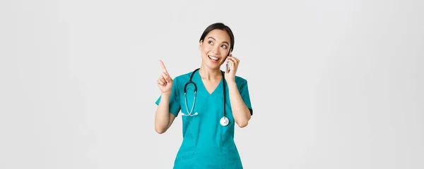 Covid-19, медицинские работники и предотвращение концепции вируса. Портрет молодой симпатичной азиатки-врача, медсестры в униформе, делающей онлайн заказ, разговаривающей по телефону и указывающей на левый верхний угол, улыбающейся — стоковое фото