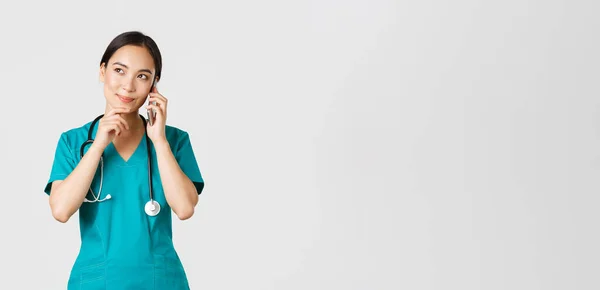 Covid-19, egészségügyi dolgozók és a vírus fogalmának megelőzése. Mosolygó ázsiai doktornő portréja, gyakornok köpenyben beszél telefonon és figyelmes, gondolkodó vagy választó. — Stock Fotó