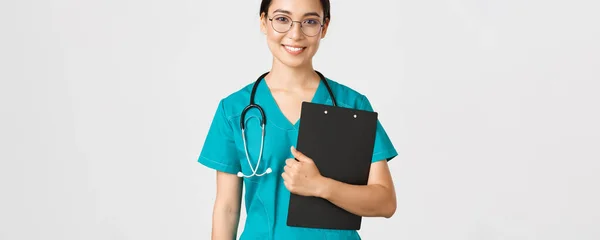 Ковид-19, коронавирусная болезнь, концепция медицинских работников. Улыбающаяся уверенная азиатка медсестра, врач в очках и халатах держит планшет, делает осмотр, белый фон — стоковое фото