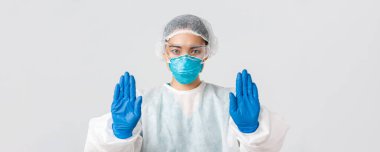 Covid-19, koronavirüs hastalığı, sağlık çalışanları konsepti. Ciddi görünümlü endişeli Asyalı kadın doktorlara yakın çekim. Kişisel koruma ekipmanlarında dur hareketi göster, tehlikeli bölgeye girmeni yasakla.