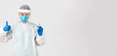Covid-19, koronavirüs hastalığı, sağlık çalışanları konsepti. Kişisel koruma ekipmanlarında kendine güvenen ciddi Asyalı kadın doktor, baş parmağını kaldır ve aşı ile şırıngayı tut, beyaz arka plan