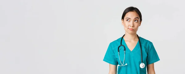 Pracownicy służby zdrowia, zapobieganie wirusom, koncepcja kampanii kwarantanny. Przemyślany azjatycki lekarz, pielęgniarka w fartuchu patrząc w lewy górny róg i uśmiech niepewny, myślenie, białe tło — Zdjęcie stockowe