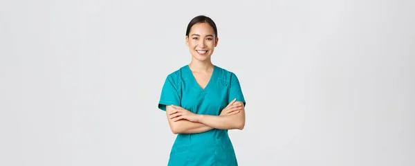 Covid-19, sağlık çalışanları, salgın konsepti. Kendinden emin gülümseyen Asyalı doktor, önlüklü kadın hemşire kararlı duruyor, beyaz arka plan üzerinde çapraz kollar göğsü. Doktor klinikte vardiyaya hazır. — Stok fotoğraf