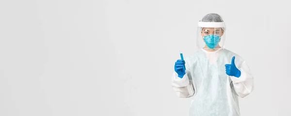Covid-19, onemocnění koronavirem, koncept zdravotnických pracovníků. Sebevědomý vážné asijské žena lékař, tech laborant v osobní ochranné prostředky show thumb-up and hold test-tube with vaccine — Stock fotografie
