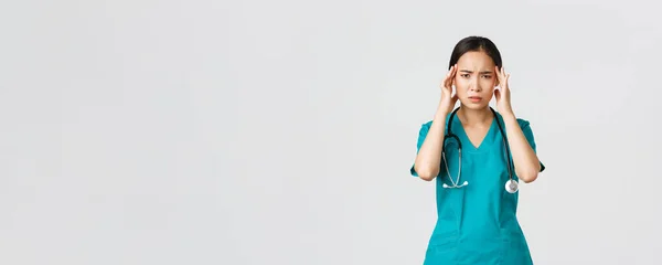 Ковід-19, медичні працівники, пандемія. Перевантажений і виснажений азіатський лікар-жінка, лікар відчуває себе хворим, носить скраби, торкається голови, скаржиться на головний біль або високу температуру — стокове фото