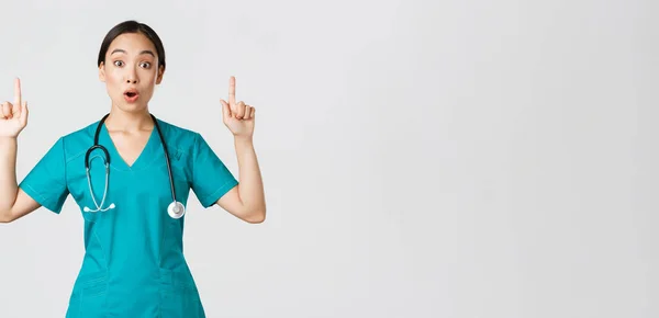 Covid-19, pracownicy służby zdrowia, koncepcja pandemii. Zaskoczona i zachwycona azjatycka pielęgniarka, lekarka w fartuchu zadająca ciekawe promo, wskazująca palcem w górę, pokazująca reklamę kliniki — Zdjęcie stockowe