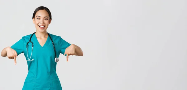 Covid-19, Gesundheitspersonal, Pandemiekonzept. Glücklich lächelnd schöne asiatische Krankenschwester, Ärztin in Peelings, die nach unten zeigt, zur Untersuchung einladend, Krankenhauswerbung zeigend, weißer Hintergrund — Stockfoto