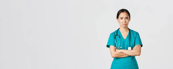Covid-19, pracownicy służby zdrowia, koncepcja pandemii. Sceptyczna i niechętna azjatycka lekarz, zmęczona pielęgniarka w fartuchu krzyżują ramiona i zmarszczki, uśmiech niezadowolony, stojące białe tło — Zdjęcie stockowe