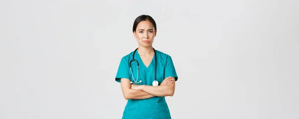 Covid-19, pracownicy służby zdrowia, koncepcja pandemii. Sceptyczna i niechętna azjatycka lekarz, zmęczona pielęgniarka w fartuchu krzyżują ramiona i zmarszczki, uśmiech niezadowolony, stojące białe tło — Zdjęcie stockowe