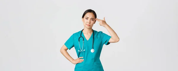 Covid-19, sjukvårdspersonal, pandemikoncept. Avtrubbad och besvärad, irriterad asiatisk kvinnlig läkare, sjuksköterska gör skott pistol gest över huvudet från tristess, vit bakgrund — Stockfoto